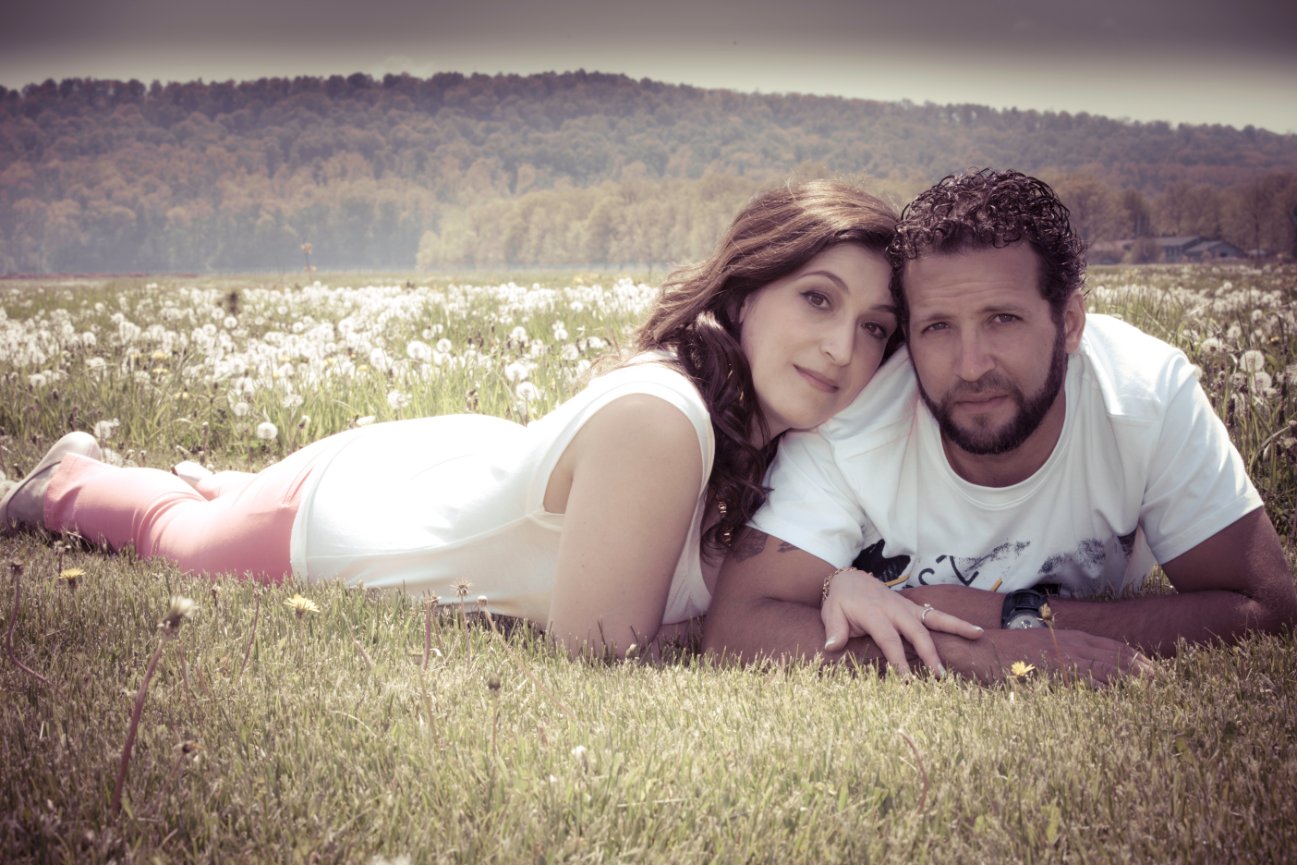 Couple lying in a field of dandelions with a forested hill in the background.