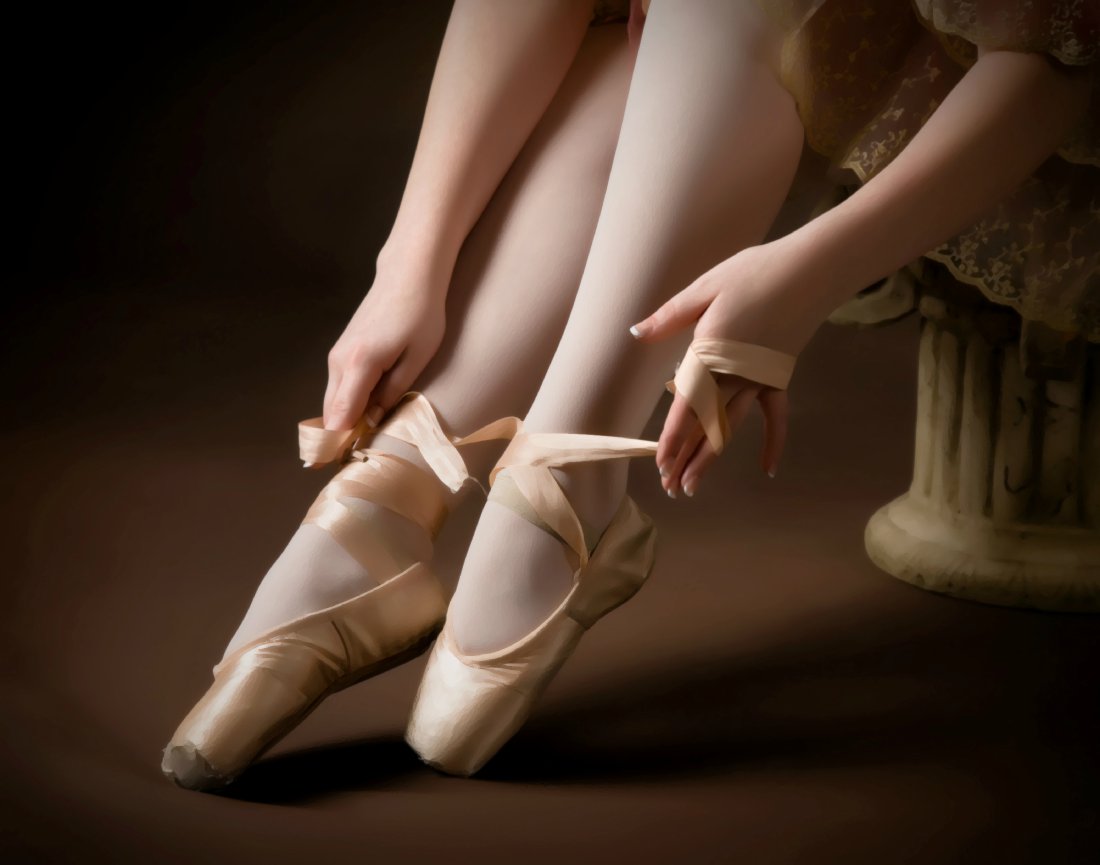 Ballerina adjusting pink pointe shoes, sitting gracefully on a stool, with focus on hands and feet.