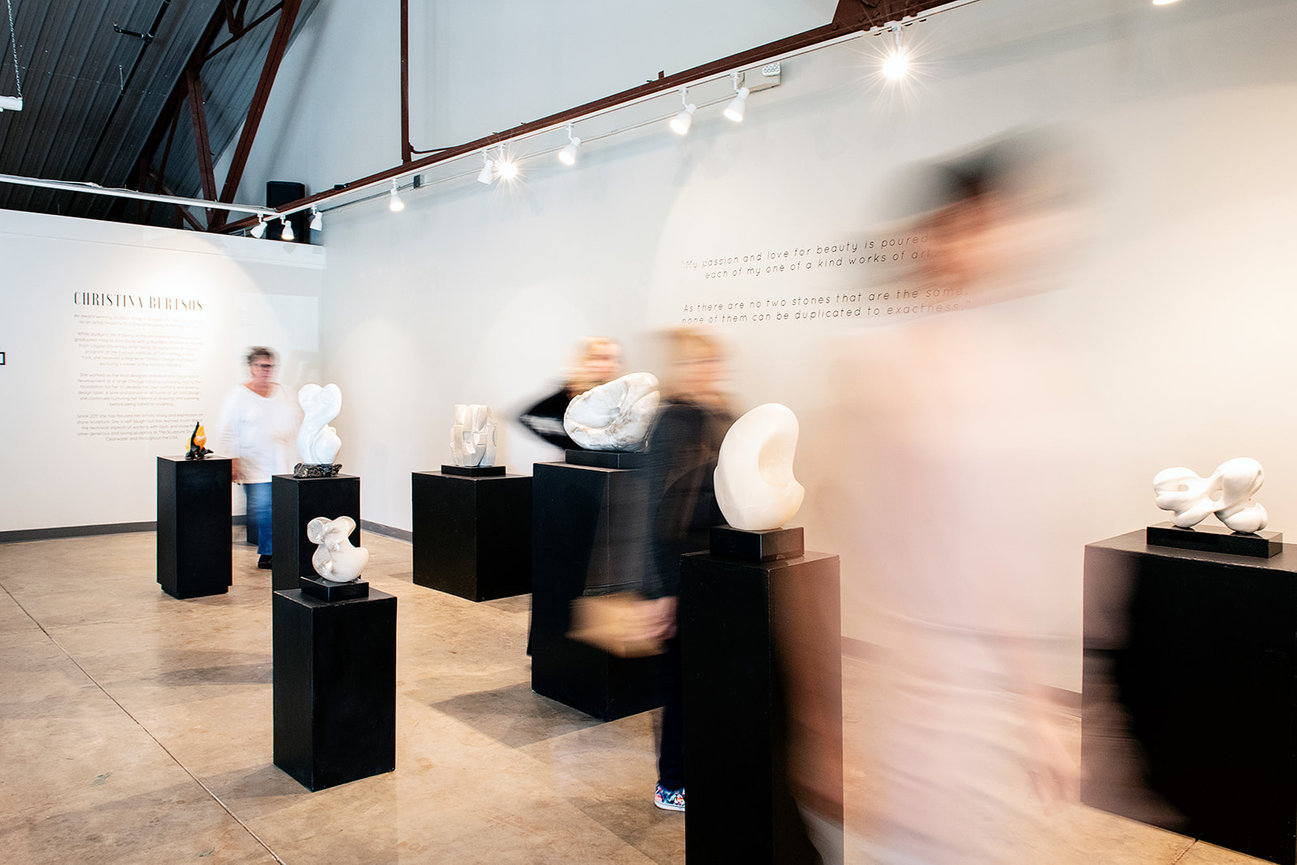 Visitors explore an art gallery featuring white marble sculptures by Christina Belkin, with blurred figures in motion.