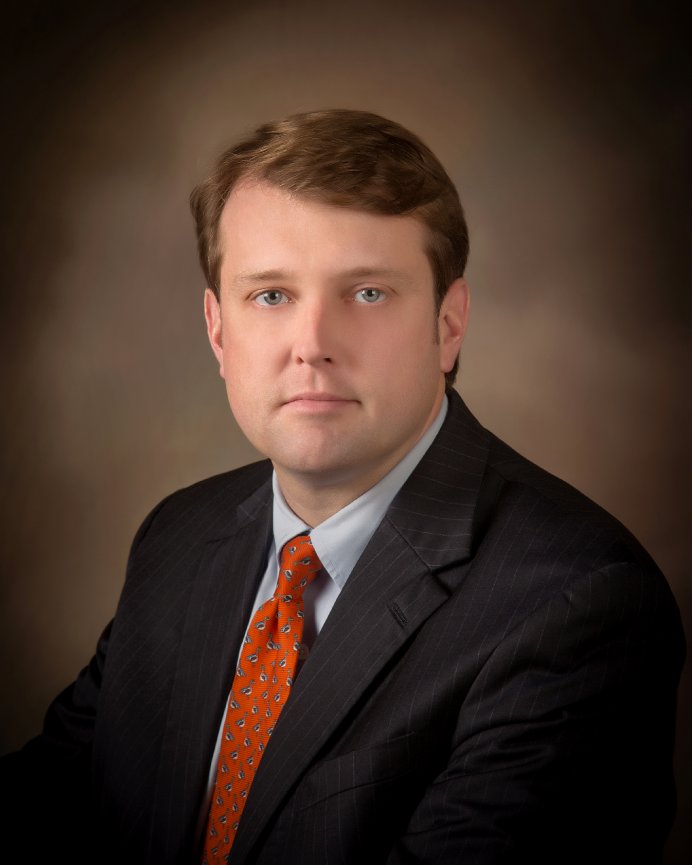A man in a suit and orange tie with a neutral expression against a brown background.