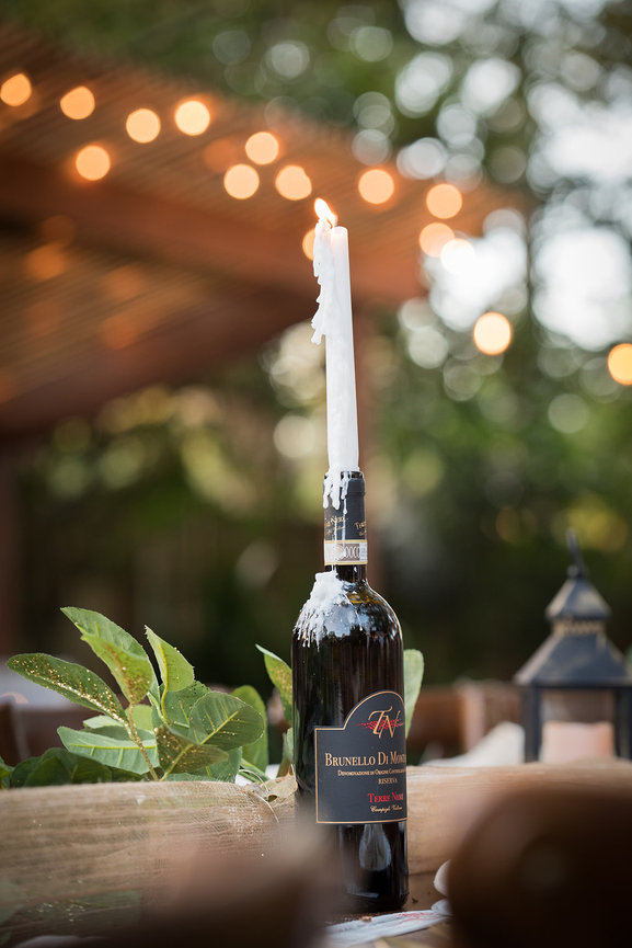 A bottle of Brunello di Montalcino wine with a lit candle on top, set against a blurred outdoor background with string lights.
