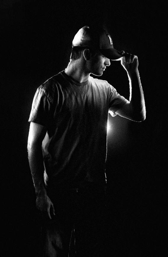 Man in a T-shirt and cap, lit dramatically from behind in a dark setting.