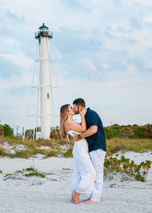 Couple kissing on a sandy beach with a lighthouse in the background.