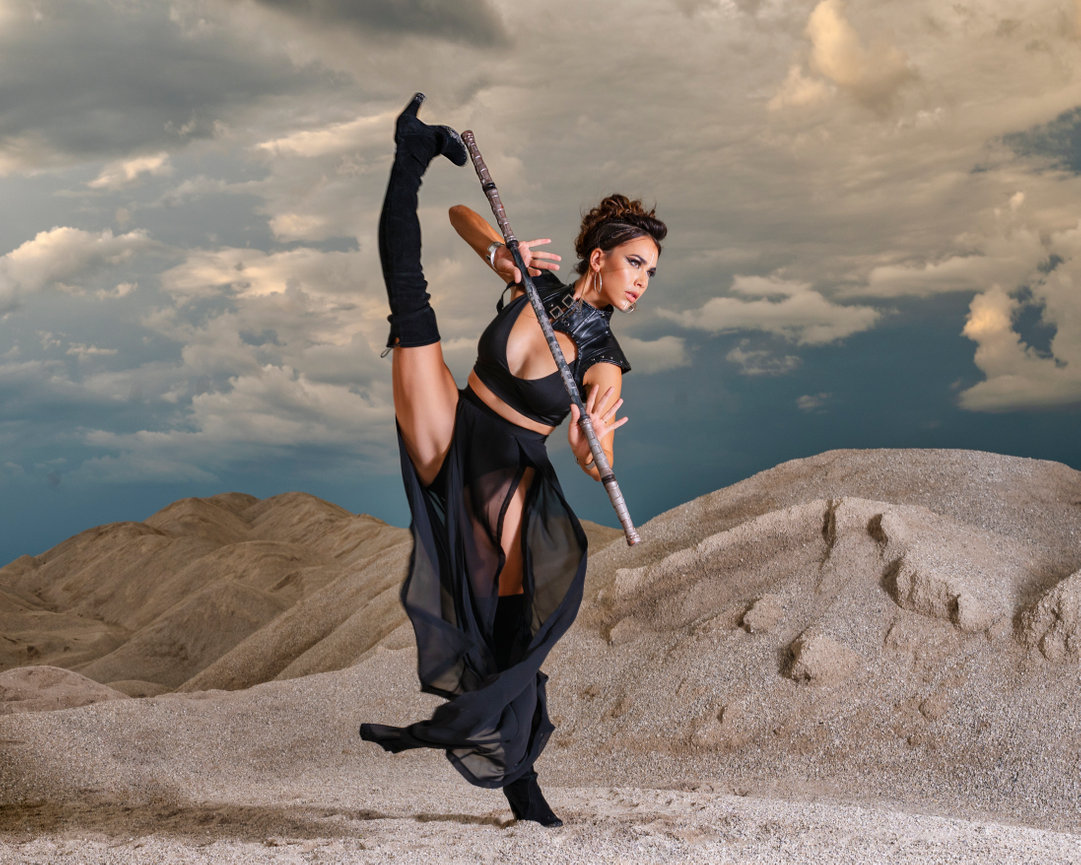 Dancer in a dynamic pose, wearing a black outfit, striking against a dramatic sky and sandy landscape.
