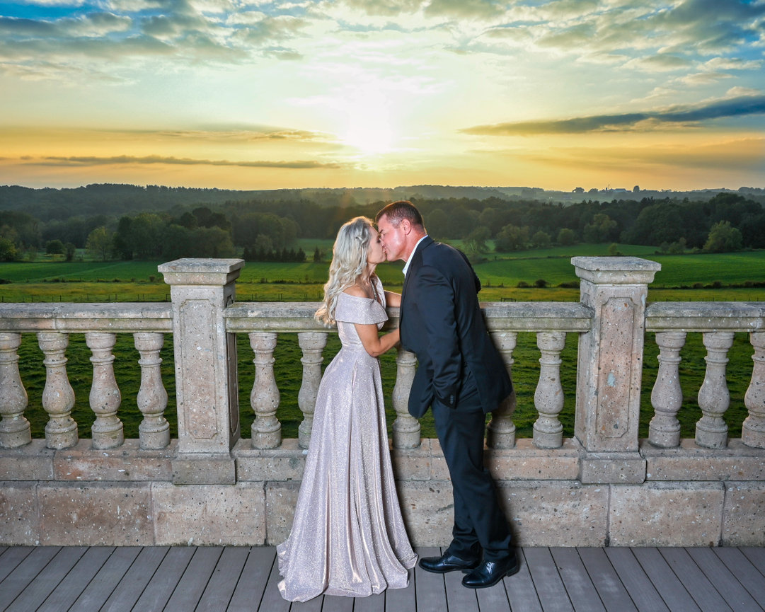 Couple in formal attire kissing at sunset on a balcony with scenic fields in the background.