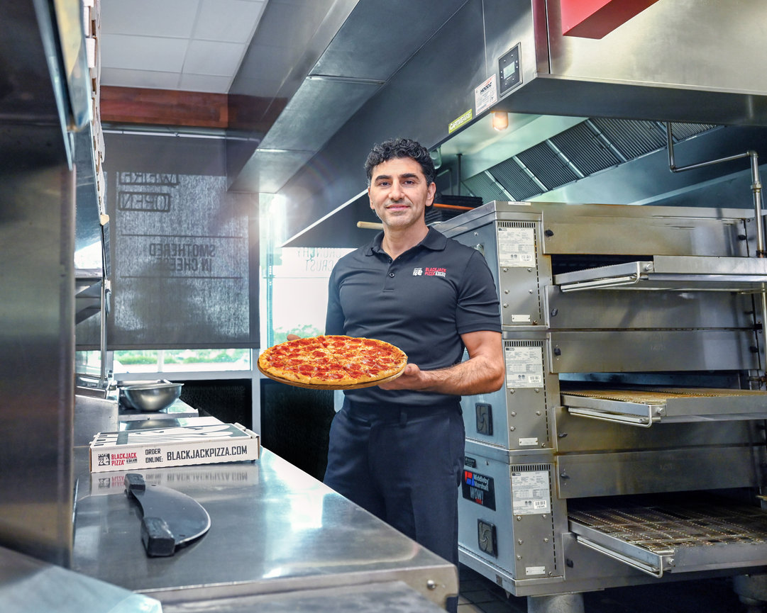 A chef holding a freshly baked pizza in a modern kitchen with ovens and stainless steel equipment.