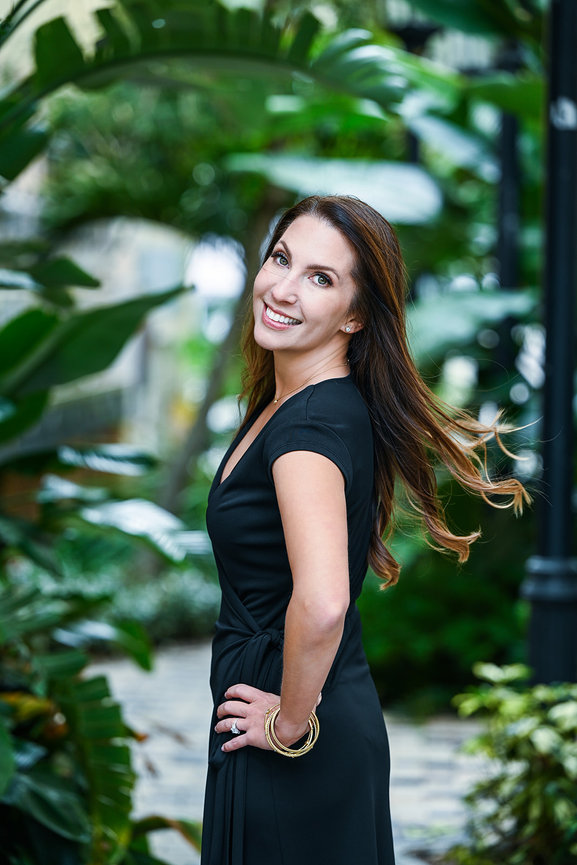Woman in a black dress smiles confidently against a lush green background of tropical plants.