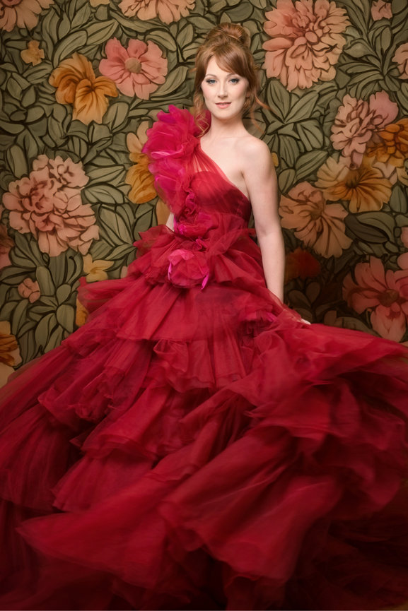 Model in a vibrant red, layered tulle gown with floral accents, posed against a floral backdrop.