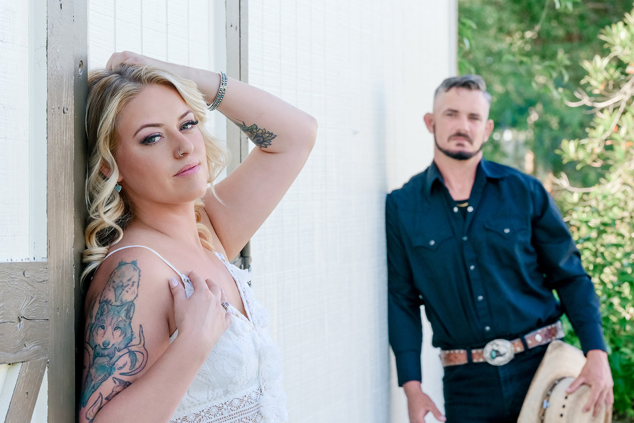 Couple posing outdoors; woman in a white dress with tattoos, man in a black shirt and cowboy hat.