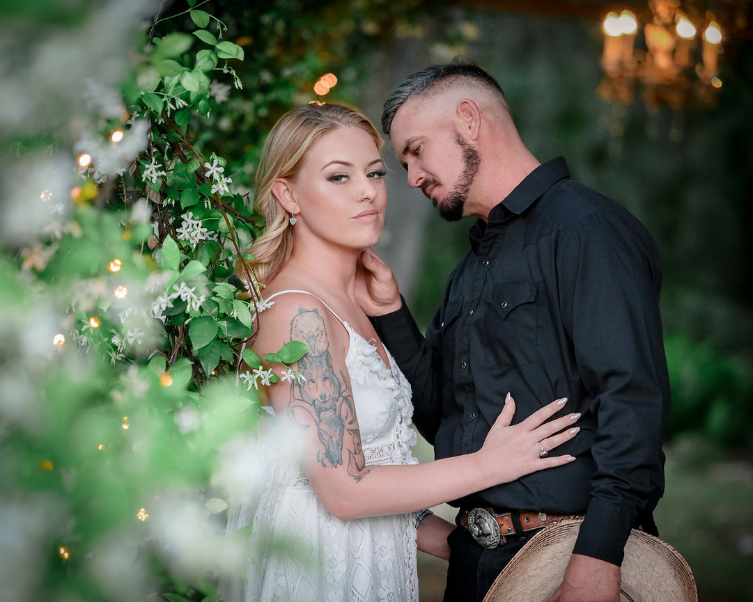 A couple poses together amidst greenery; the woman in a white dress and the man in a black shirt, creating an intimate moment.