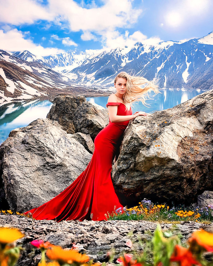 Woman in a red gown poses on rocks with a mountain lake and snow-capped peaks in the background.