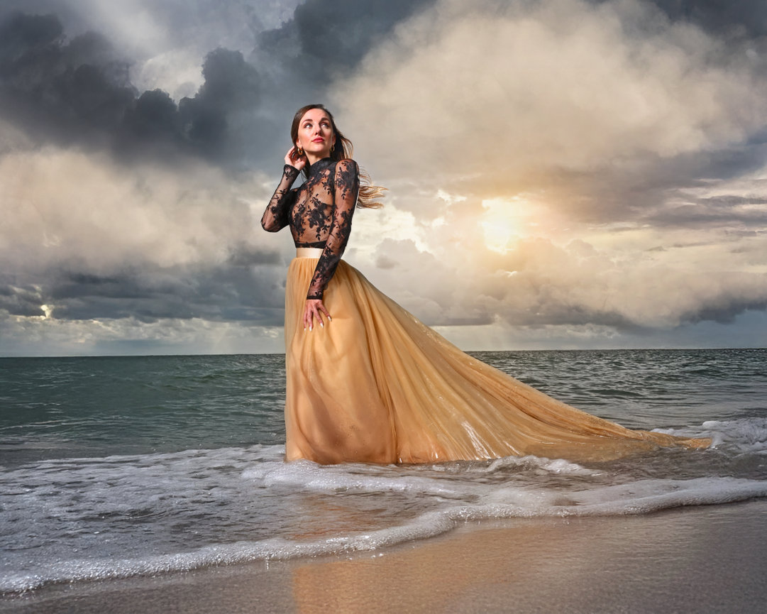 Woman in a flowing gold skirt and black lace top standing in ocean waves under moody, cloudy sky at sunset.