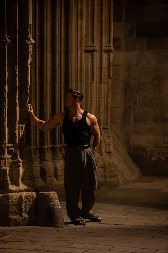 Man in a black tank top leaning against an ornate stone wall at night. Barcelona Night
