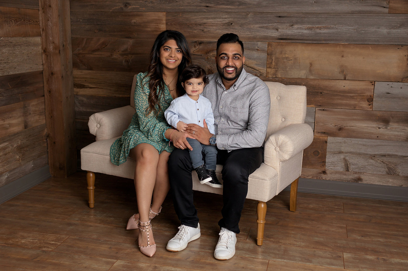A family of three sits on a beige sofa against a wooden wall, smiling and posing for a photo.