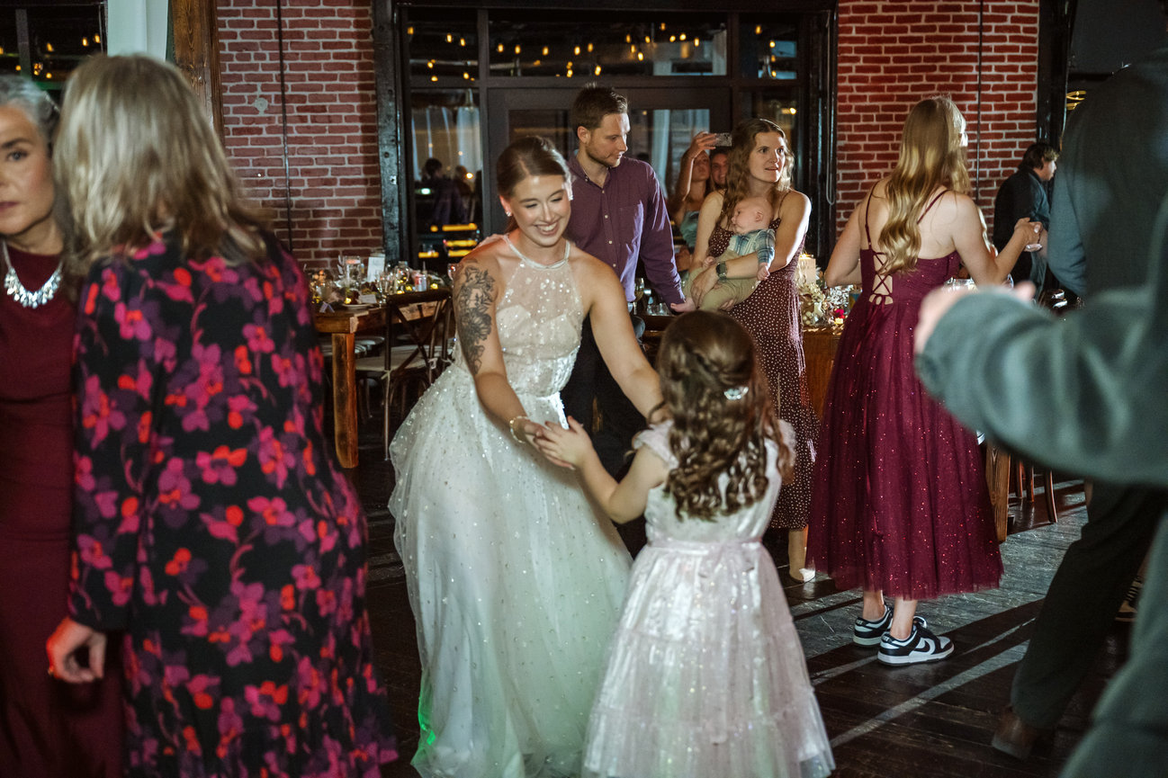 Guests dancing at a wedding reception in a brick-walled venue, with women and children in formal dresses