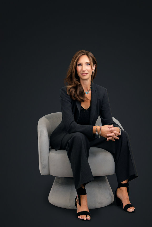 A woman in a dark suit sits confidently on a gray chair against a dark background.