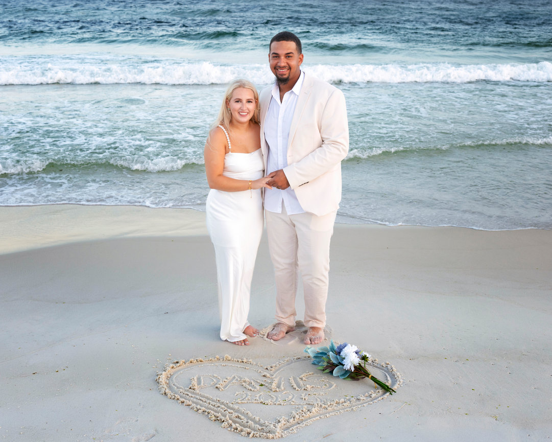 Couple holding hands at the shoreline after their Navarre Beach elopement, with a heart drawn in the sand and bouquet nearby-captured by wedding photographer Dixon Creative Images