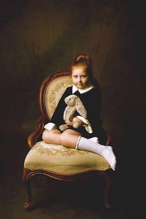 An elegant young girl with red hair in a bun sits in a Victorian ladies chair while holding a bunny lovey.