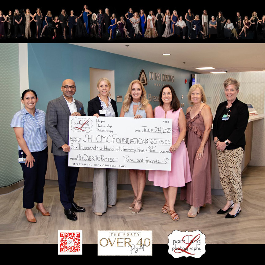 Group photo of seven adults holding a large ceremonial check at an indoor event with a crowd behind them.