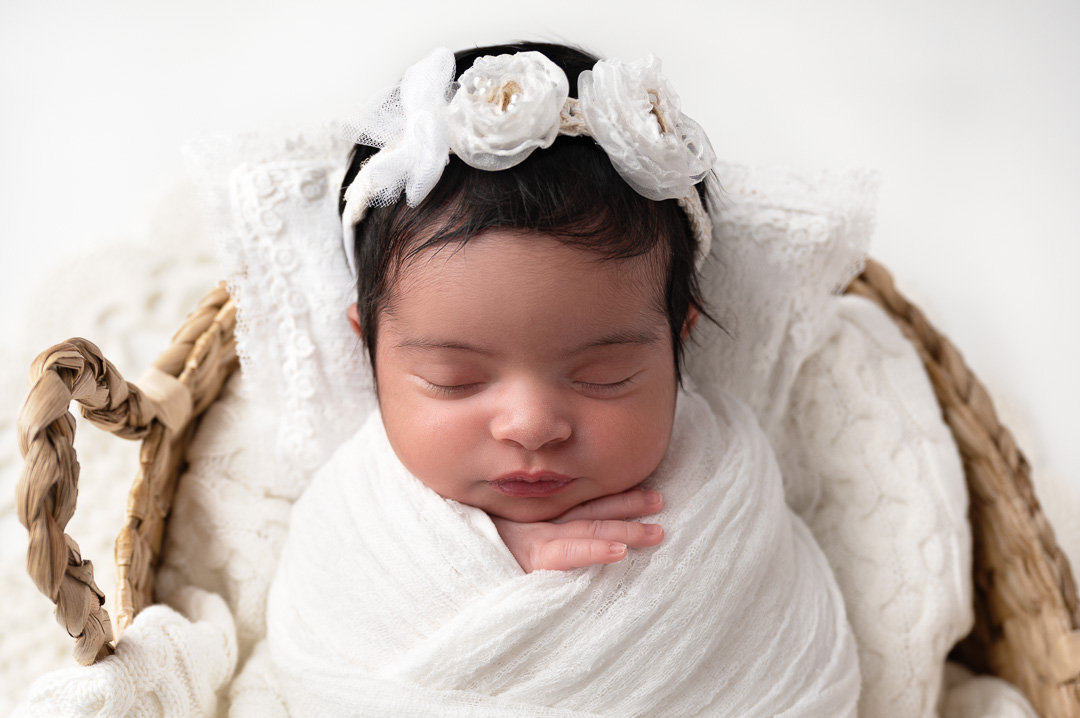 Newborn baby girl inside a basket with white headband and little hand showing