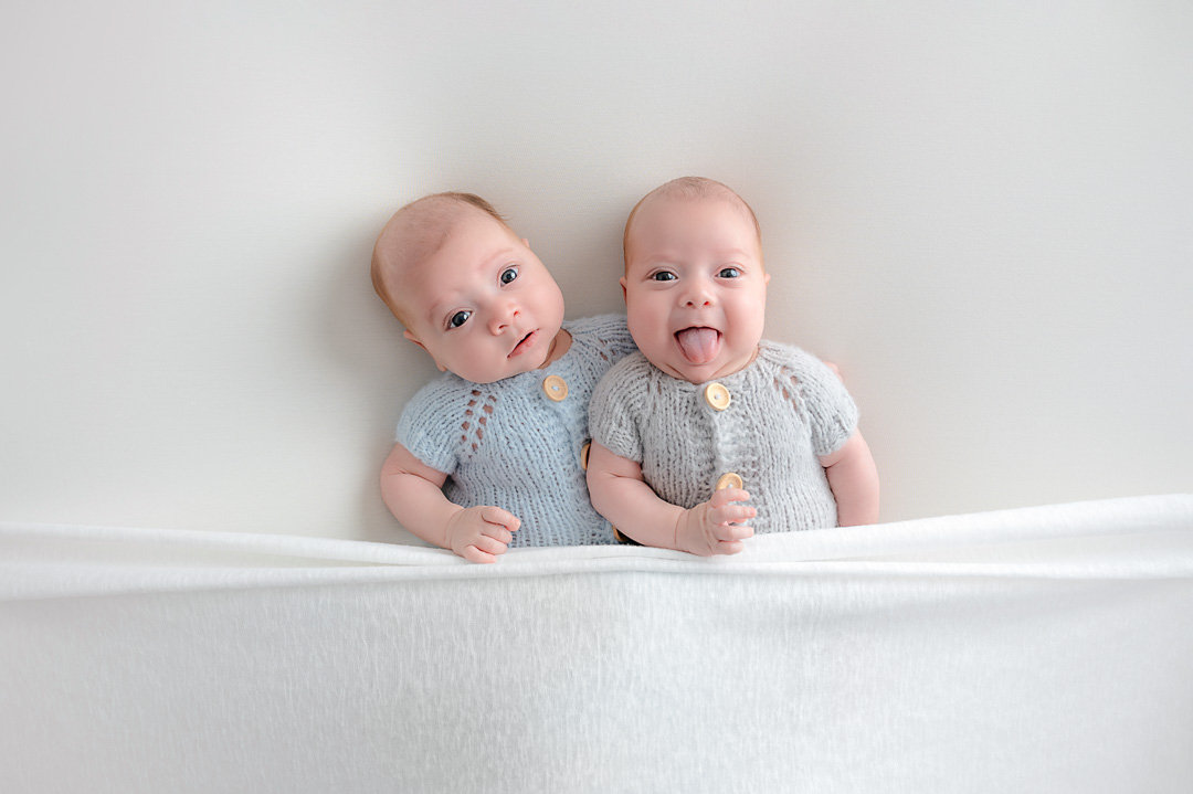Newborn twin boys next to each other, one is sticking his tongue