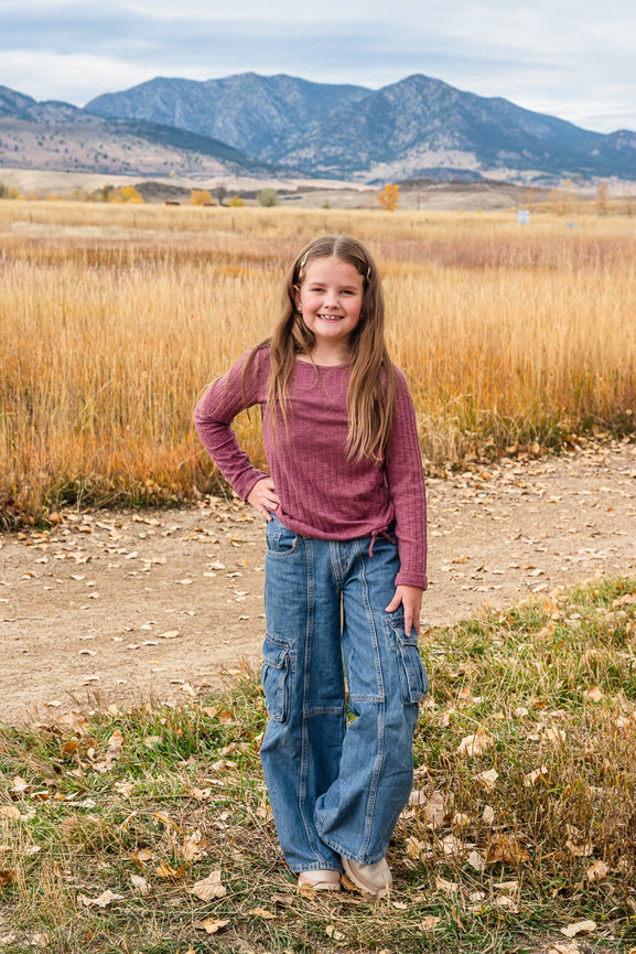 A young girl stands on a path there are dried grasses of the prairie behind her in the distance you can see the mountains.