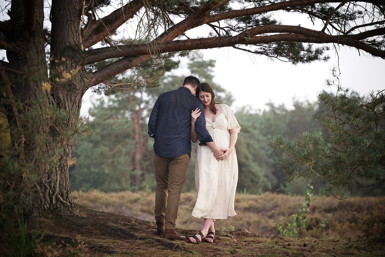 Expectant couple embracing under a tree in a field