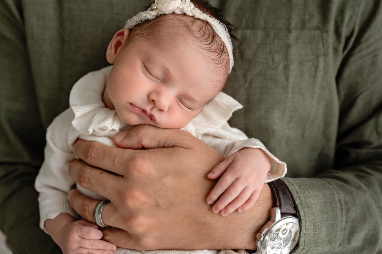 Sleeping newborn baby held in adult hands, wearing a white outfit and lace headband
