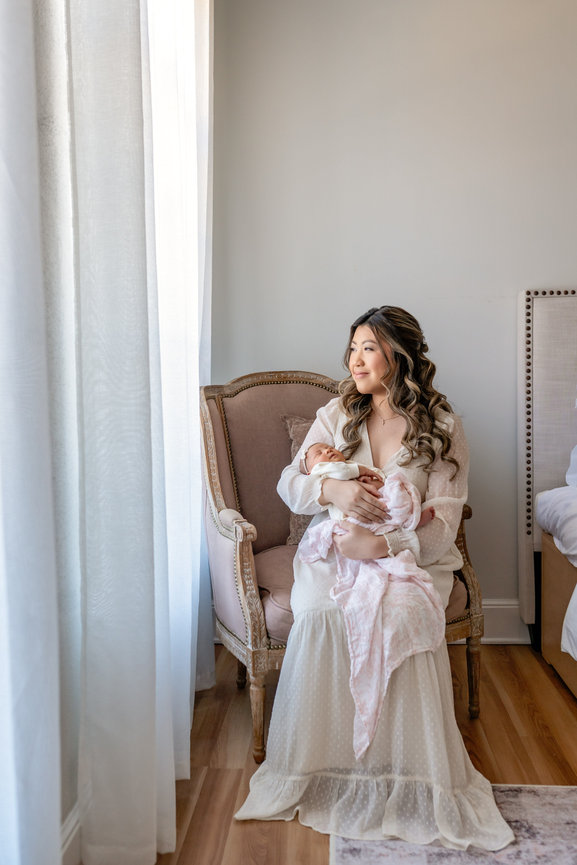 Mother holding newborn baby in a softly lit bedroom near a window.