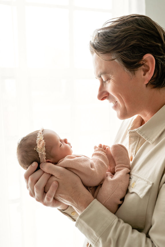 Father holding newborn baby and smiling near a bright window