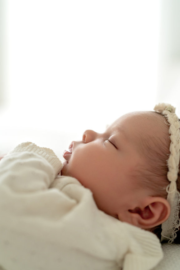 Sleeping newborn baby in white clothes with a lace headband, close-up side profile on a bright background- Melissa Madden photography