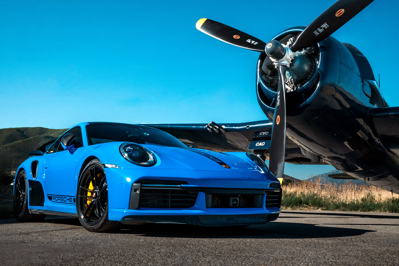 Blue Porsche parked alongside an F6F Hellcat warplane in Heber, Utah, blending luxury automotive design with historic aviation. Captured by Tre Nelson Photo.