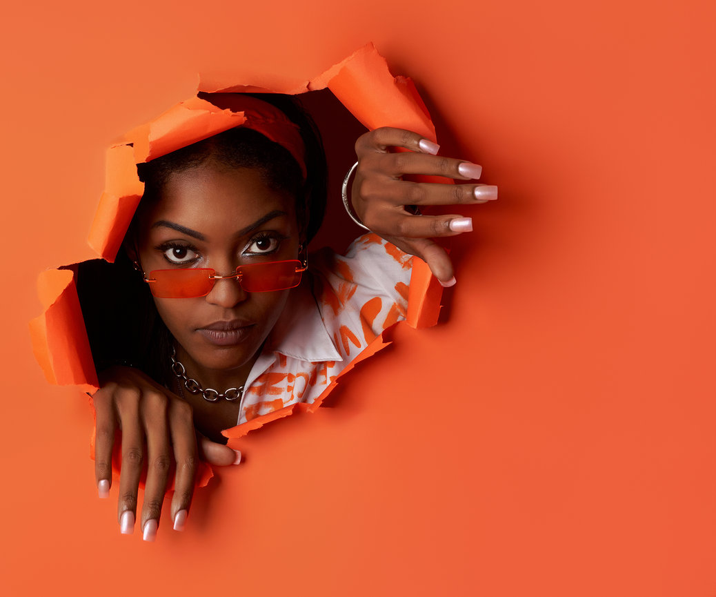 Creative personal branding portrait of a confident woman breaking through a bold orange backdrop, wearing modern fashion and orange-tinted glasses, photographed in studio by a Sherman Texas photographer