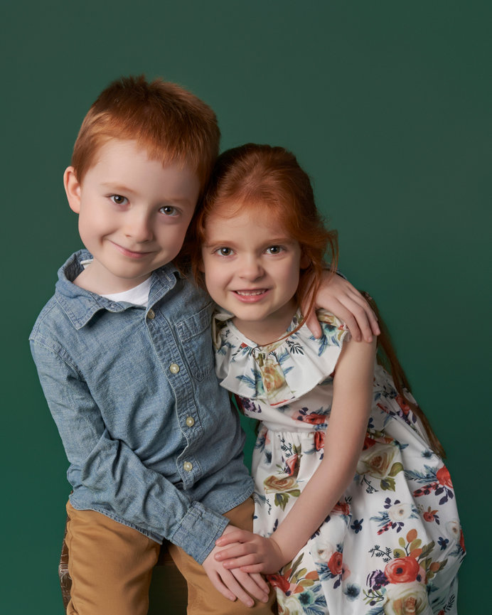Two children smiling, a boy and a girl, in colorful clothes against a green background, hugging affectionately.