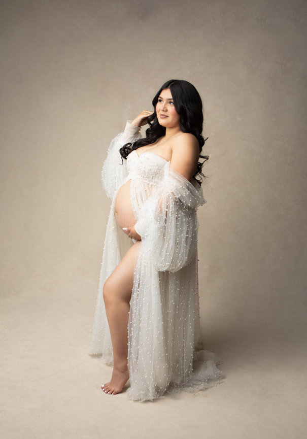 Pregnant woman in a sheer, pearl-studded gown poses against a neutral backdrop, with soft lighting.