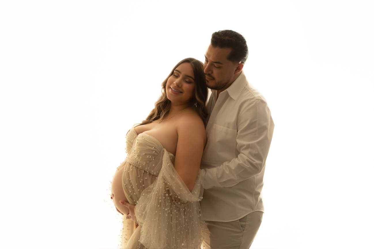 Pregnant couple embracing, woman in a tulle dress, man in a white shirt, both smiling gently.