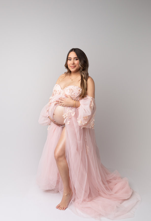 Pregnant woman in a flowing pink gown, posing barefoot against a plain backdrop.