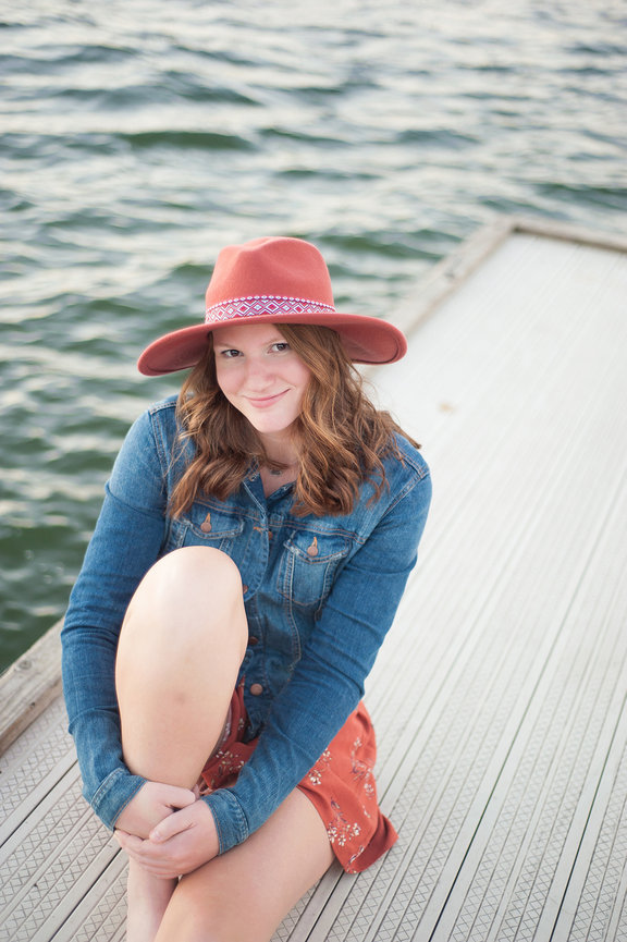 senior girl sitting on dock at Table Rock Lake in white dress, jean jacket and coral hat posing for portrait near Branson Missouri