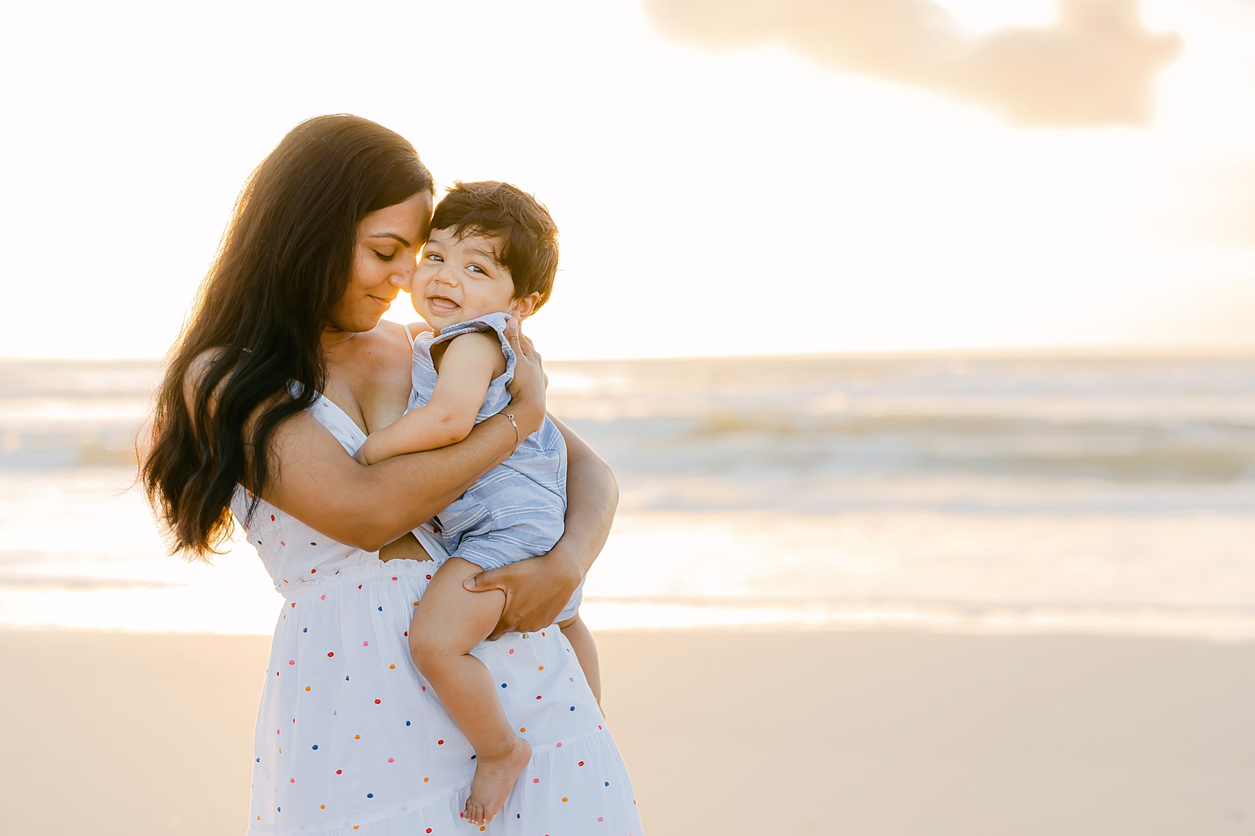 woman wearing white dress holding smiley baby at sunrise on the beach