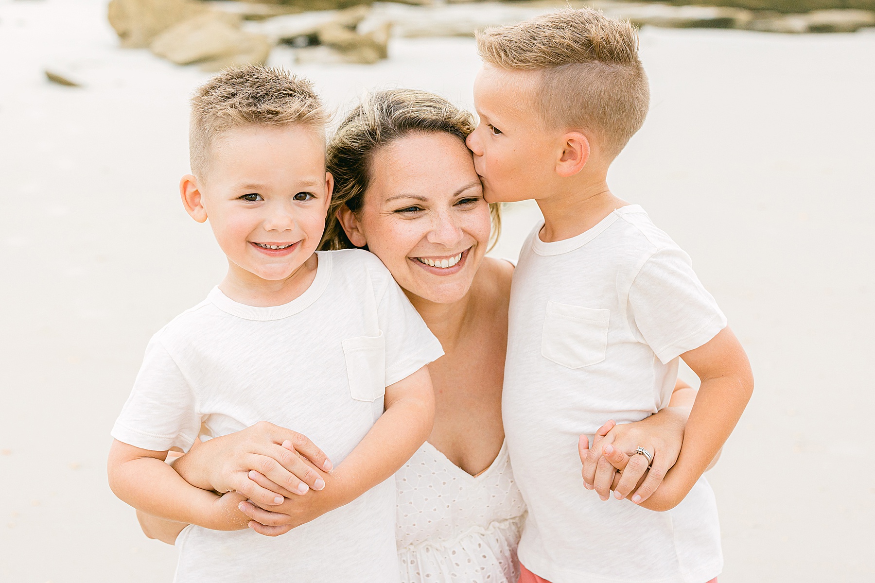 two boys kissing their mother on the beach dressed in white shirts
