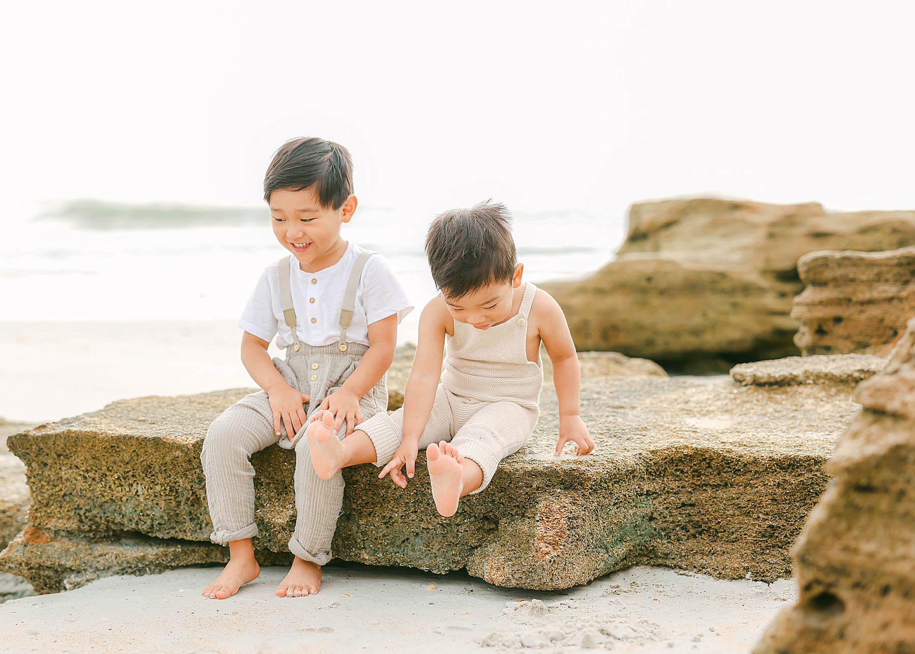 little boys sitting on rocks at the beach in overalls