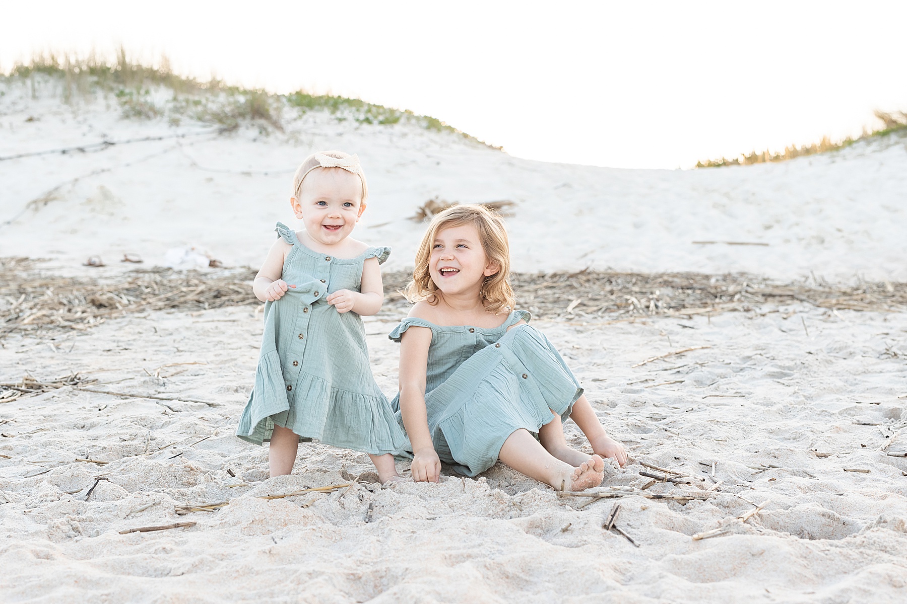 little girls in green dresses sitting on the sand on the beach laughing with each other