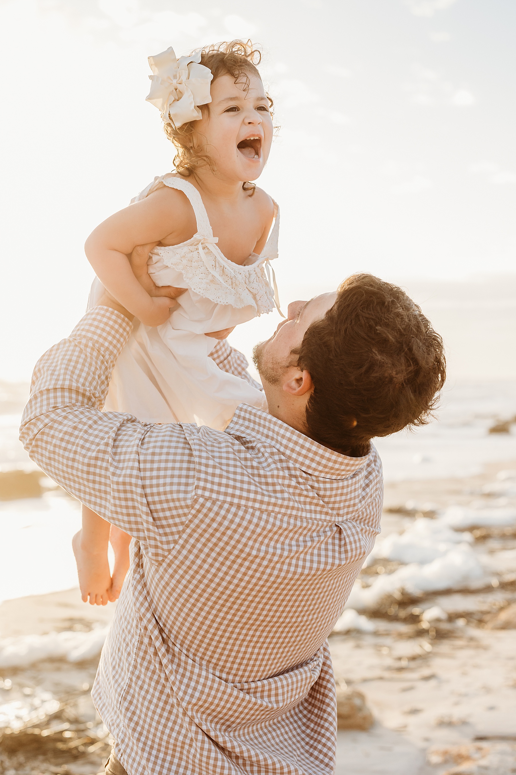 little girl being held in the air by man on the beach at sunrise laughing