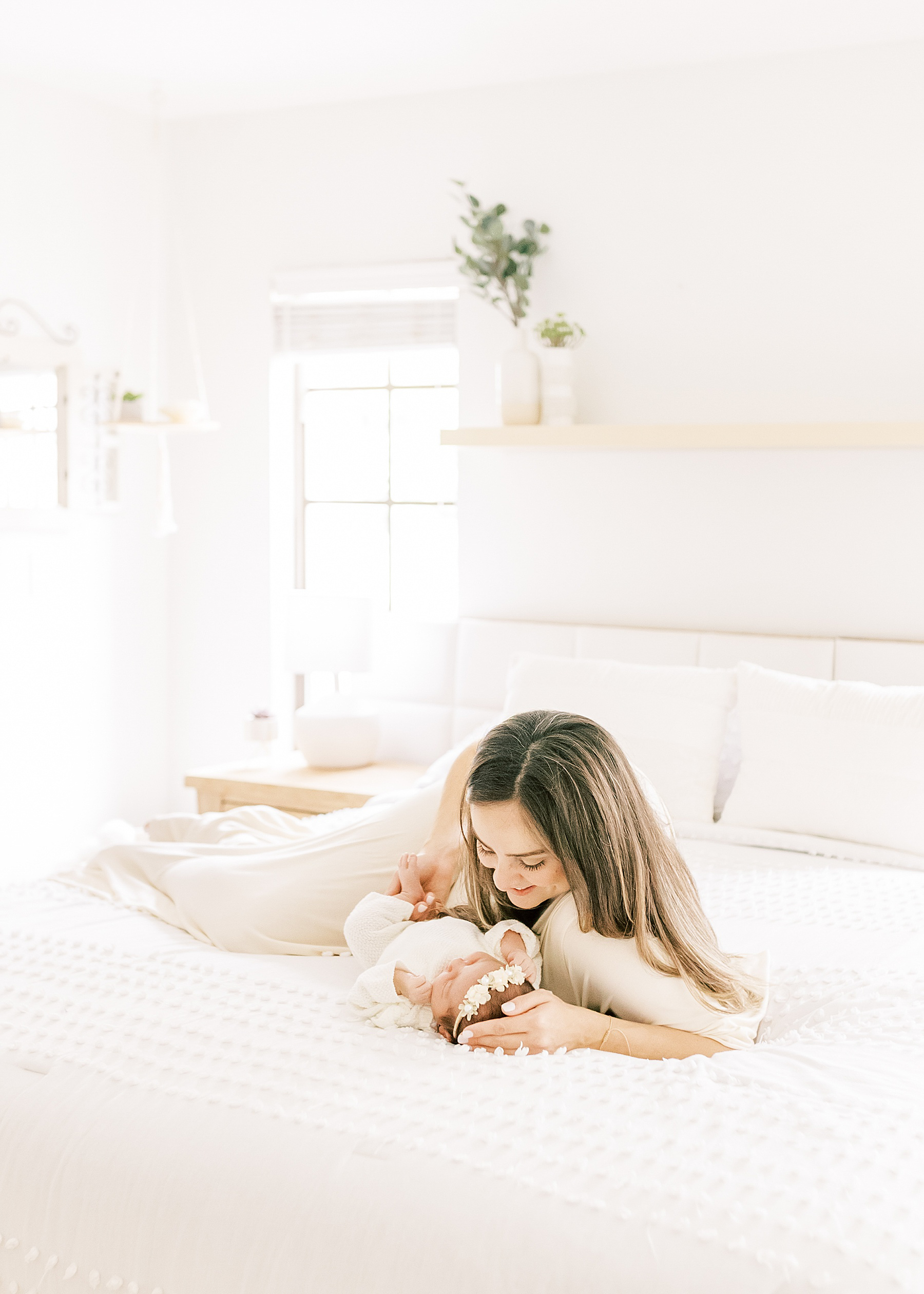 woman wearing cream dress sitting on bed with white bedding holding newborn baby