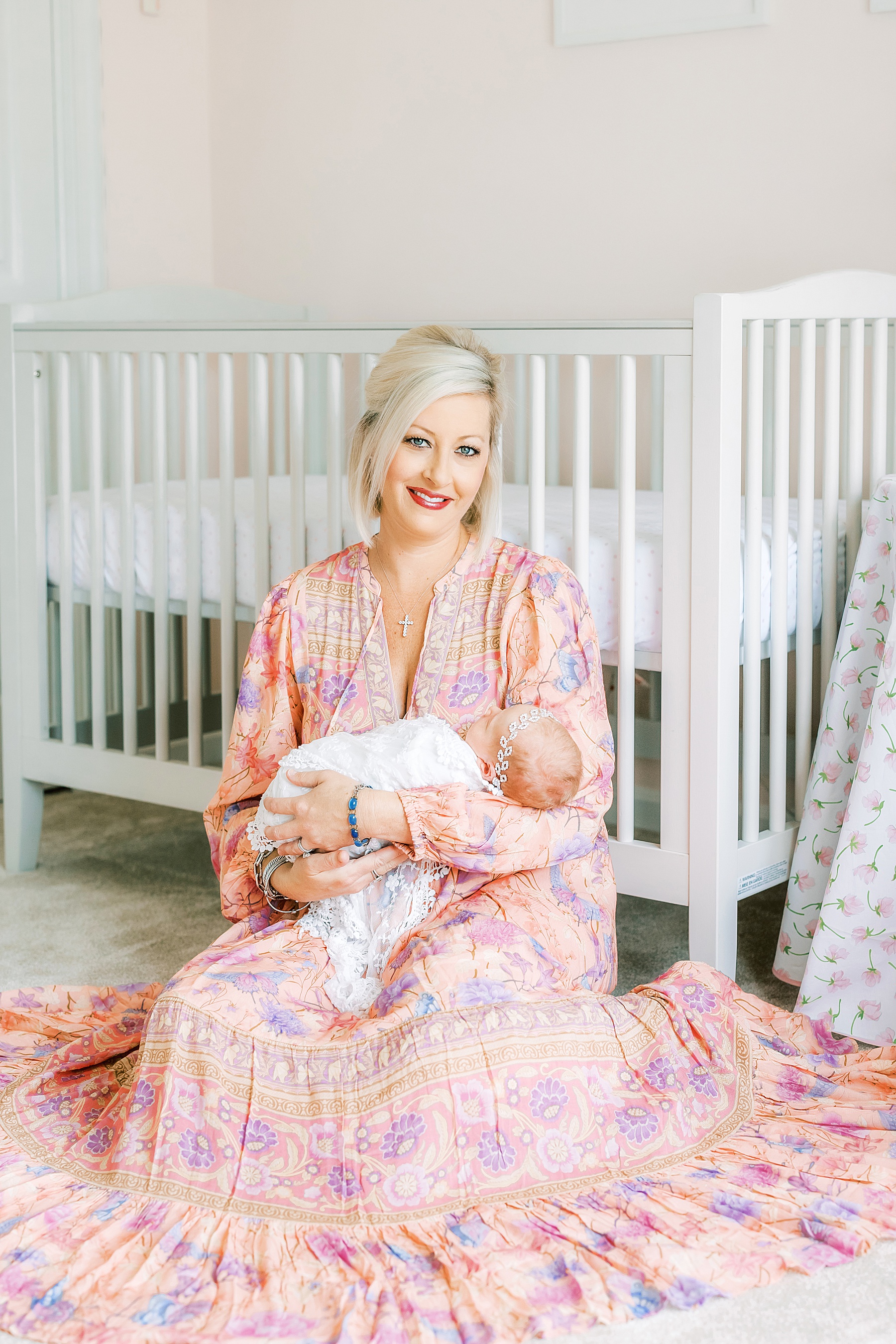 woman in pink dress holding newborn baby girl by white crib in nursery