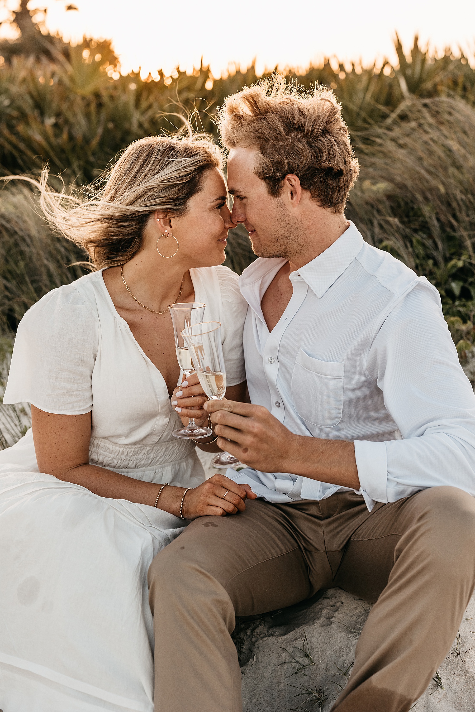 blond haired woman in long white dress snuggling man in khaki pants on the sand holding champagne