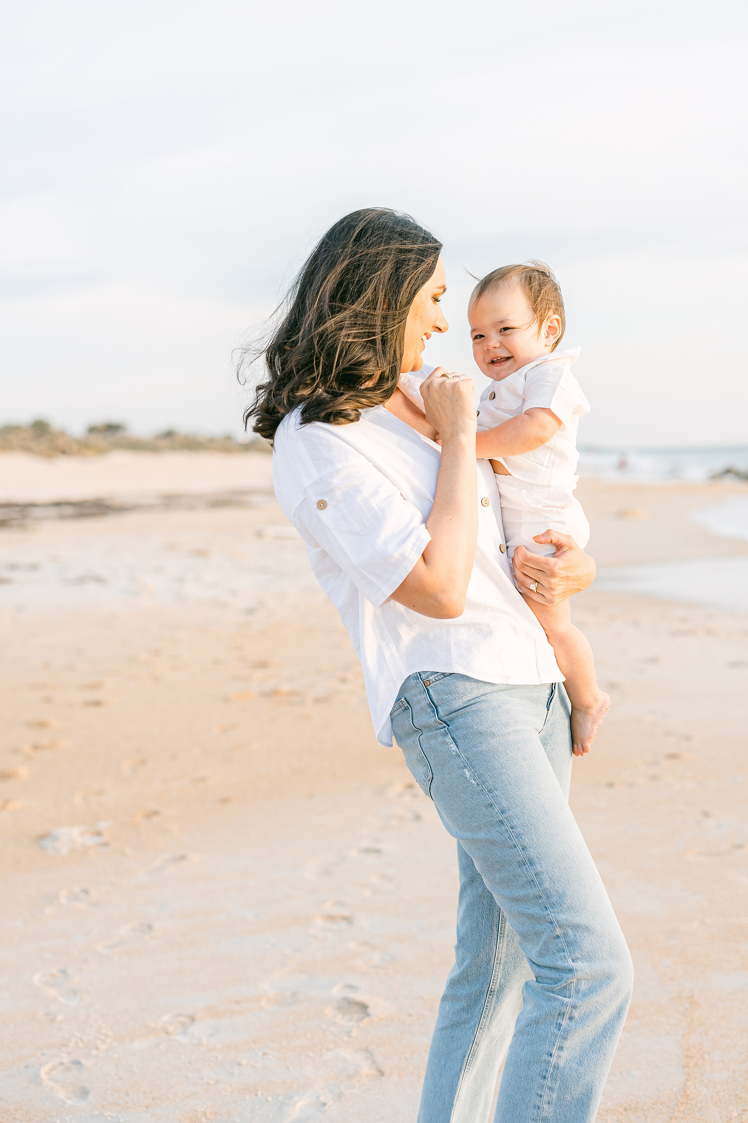 woman wearing jeans and white shirt holding baby boy on the beach at sunrise