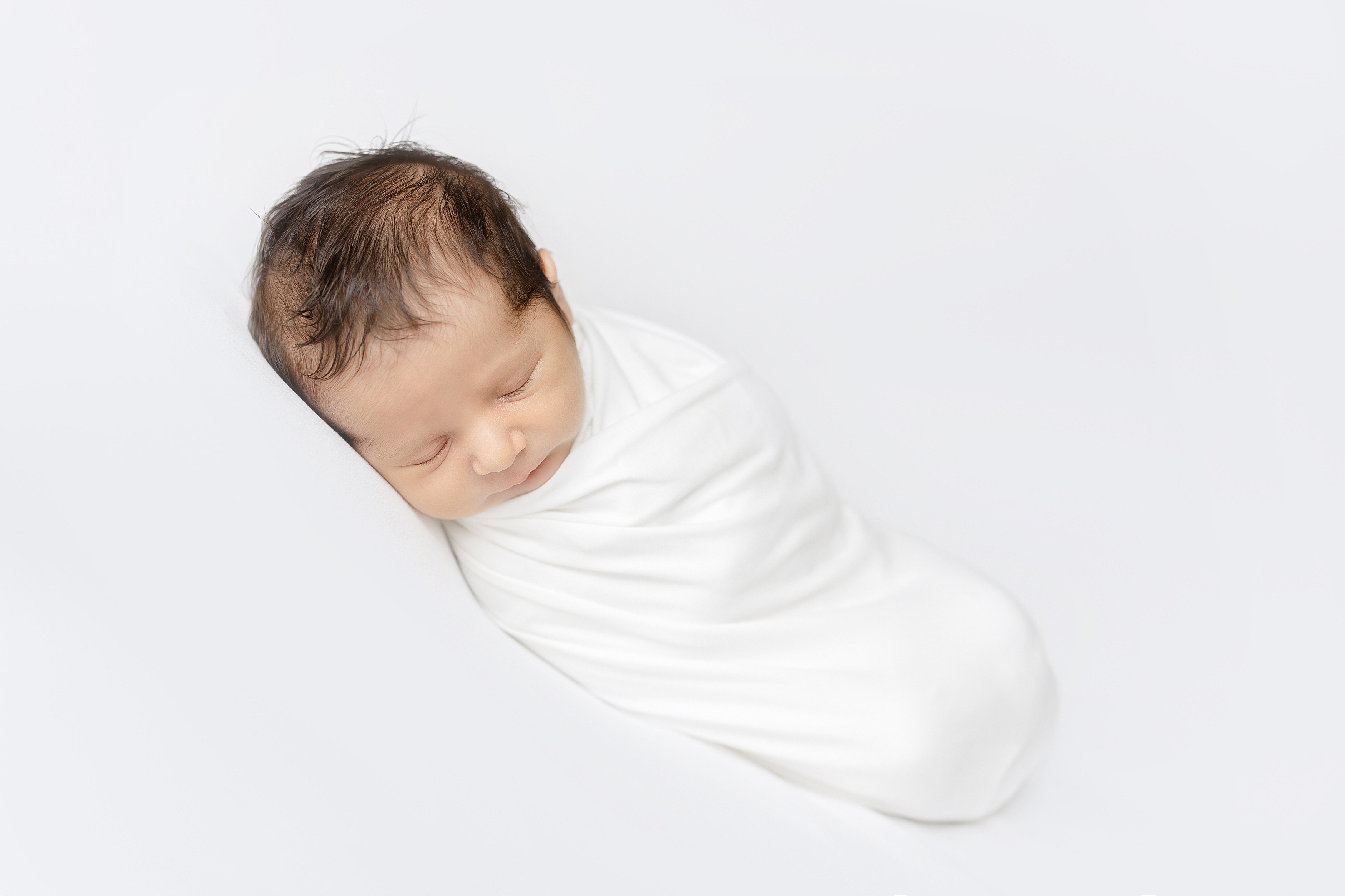 Posed newborn portrait of baby boy wrapped in white swaddle on white blanket.