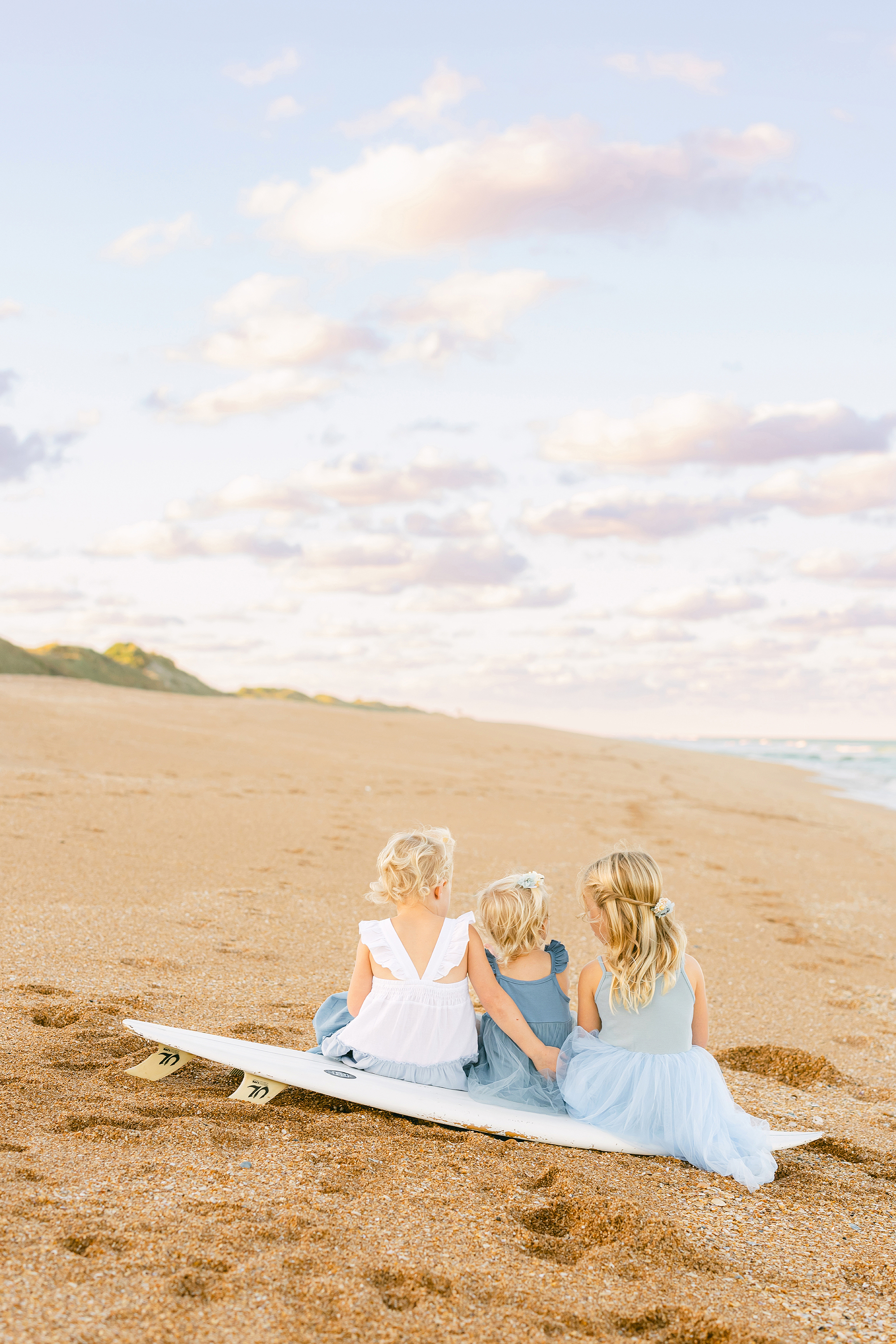 Three little girls in blue and white dresses sitting on a surfboard on the beach at sunset.