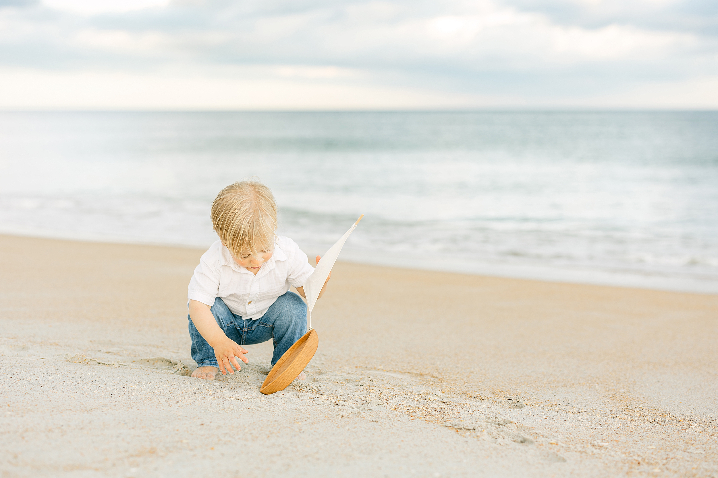 A blonde baby boy wearing jeans and a white linen shirt holds a wooden toy boat on the beach at Guana State Park.